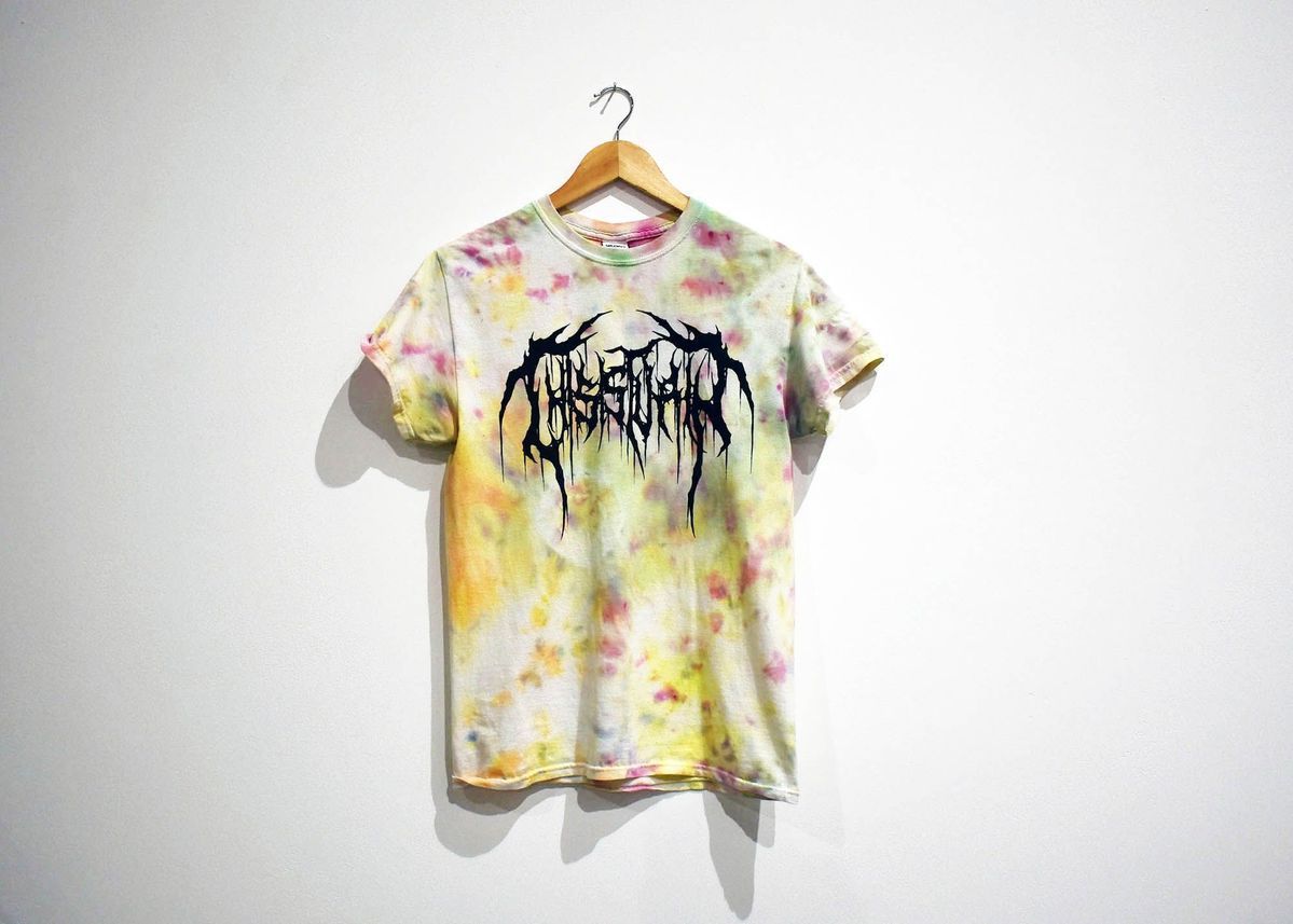 A tie-dye T-shirt with abstract lettering hangs on a wooden hanger against a plain white wall.