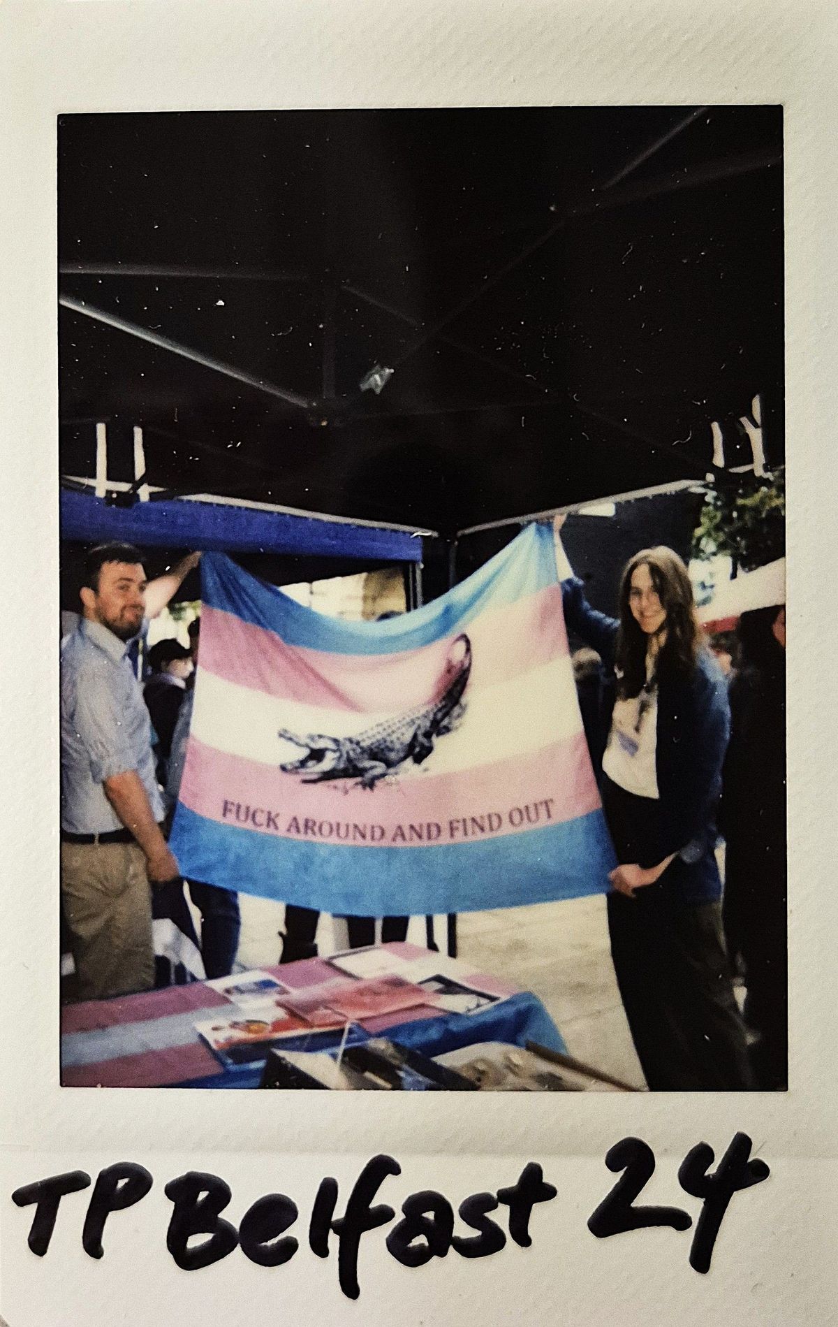 Two individuals are holding a colorful flag with text, standing at an event with tables covered in materials.
