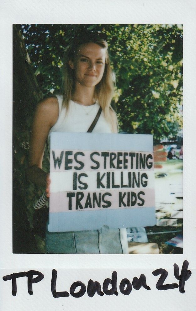 A person is holding a protest sign with bold text, standing outdoors near trees in bright daylight.