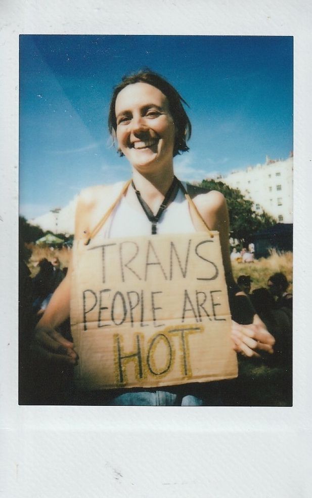 A smiling person outdoors holds a sign reading "TRANS PEOPLE ARE HOT" against a sunny blue sky.