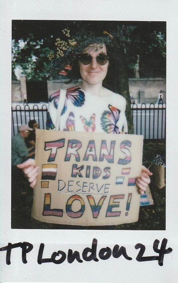 A person wearing sunglasses and a floral crown holds a colorful sign saying, "Trans kids deserve love!"
