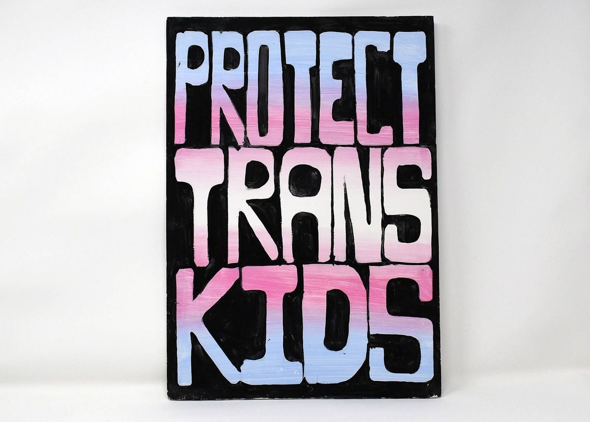 A placard which is painted black then has 'Protect Trans Kids' in large bold font in trans flag colours.