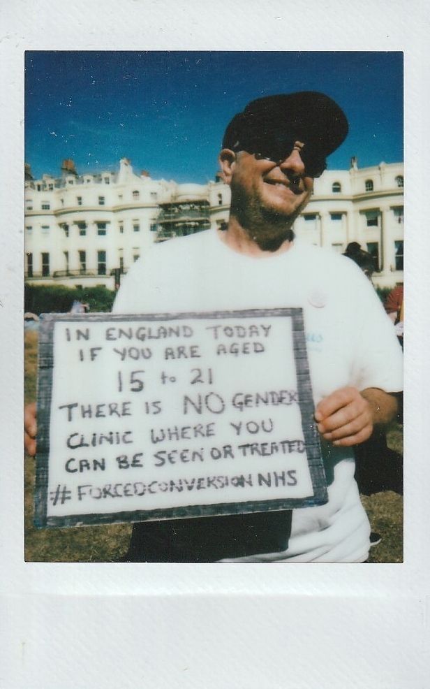A person smiling outdoors holds a sign which says "In England today if you are aged 15 to 21 there is NO gender clinic where you can be seen or treated #FORCEDCONVERSIONNHS".