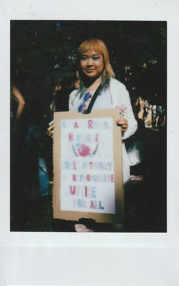 A person holds a sign which has writing on it in pink and blue but is too blurry to make out.