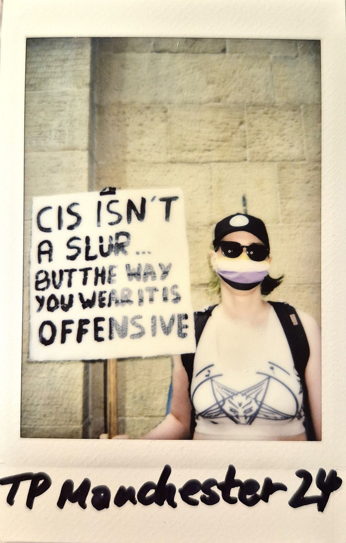 A person wearing sunglasses and a mask holds a sign reading, "Cis isn't a slur but the way you wear it is offensive".
