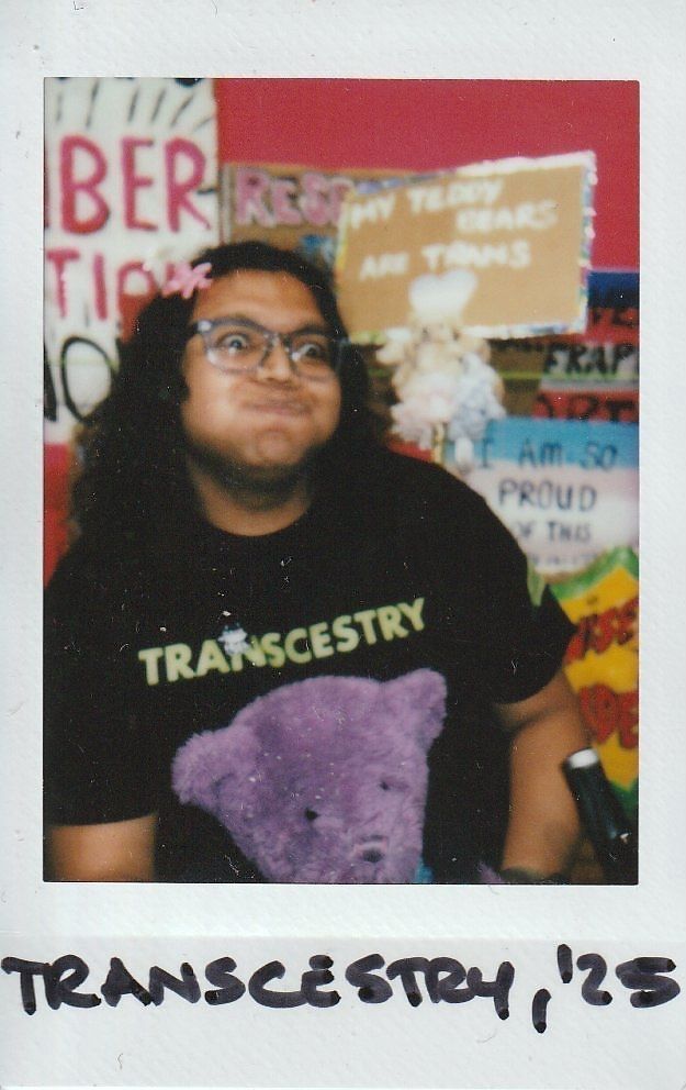 A person wearing a "TRANSCESTRY" shirt poses playfully, surrounded by colorful signs.