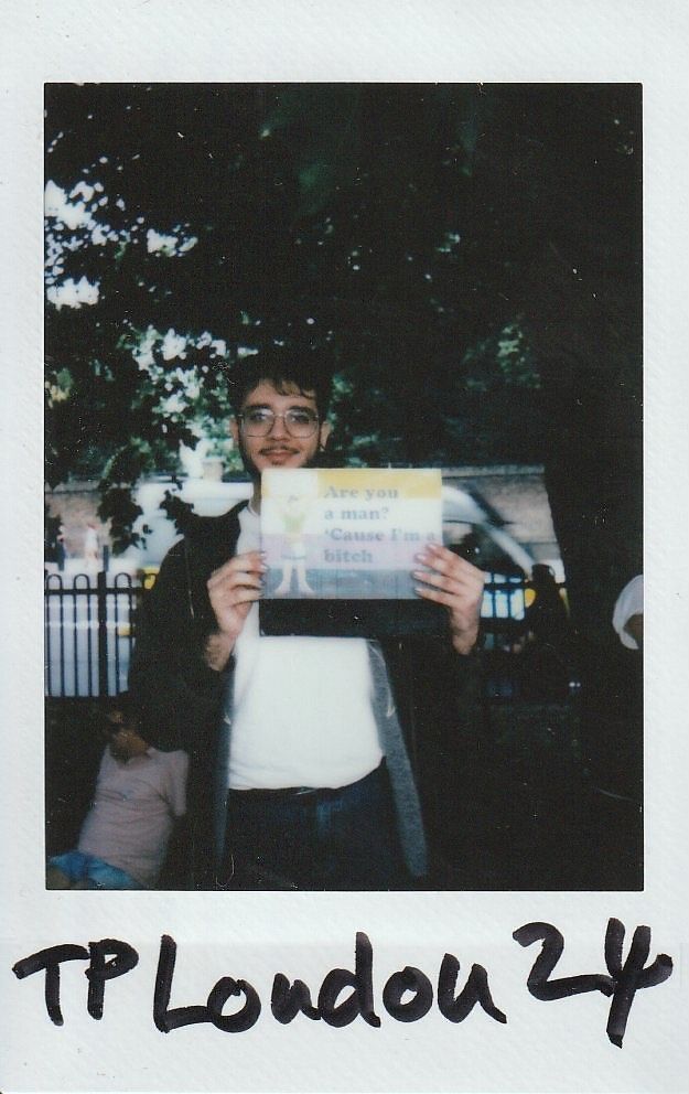 A person holding a humorous sign stands in front of a tree in a park setting.