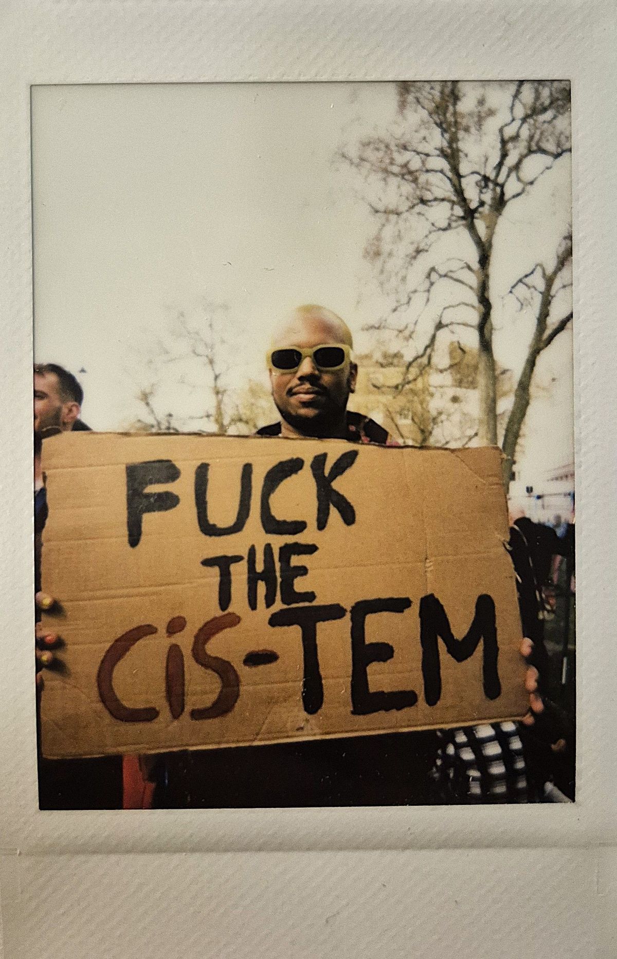 A person wearing sunglasses holds a protest sign that reads, “FUCK THE CIS-TEM".
