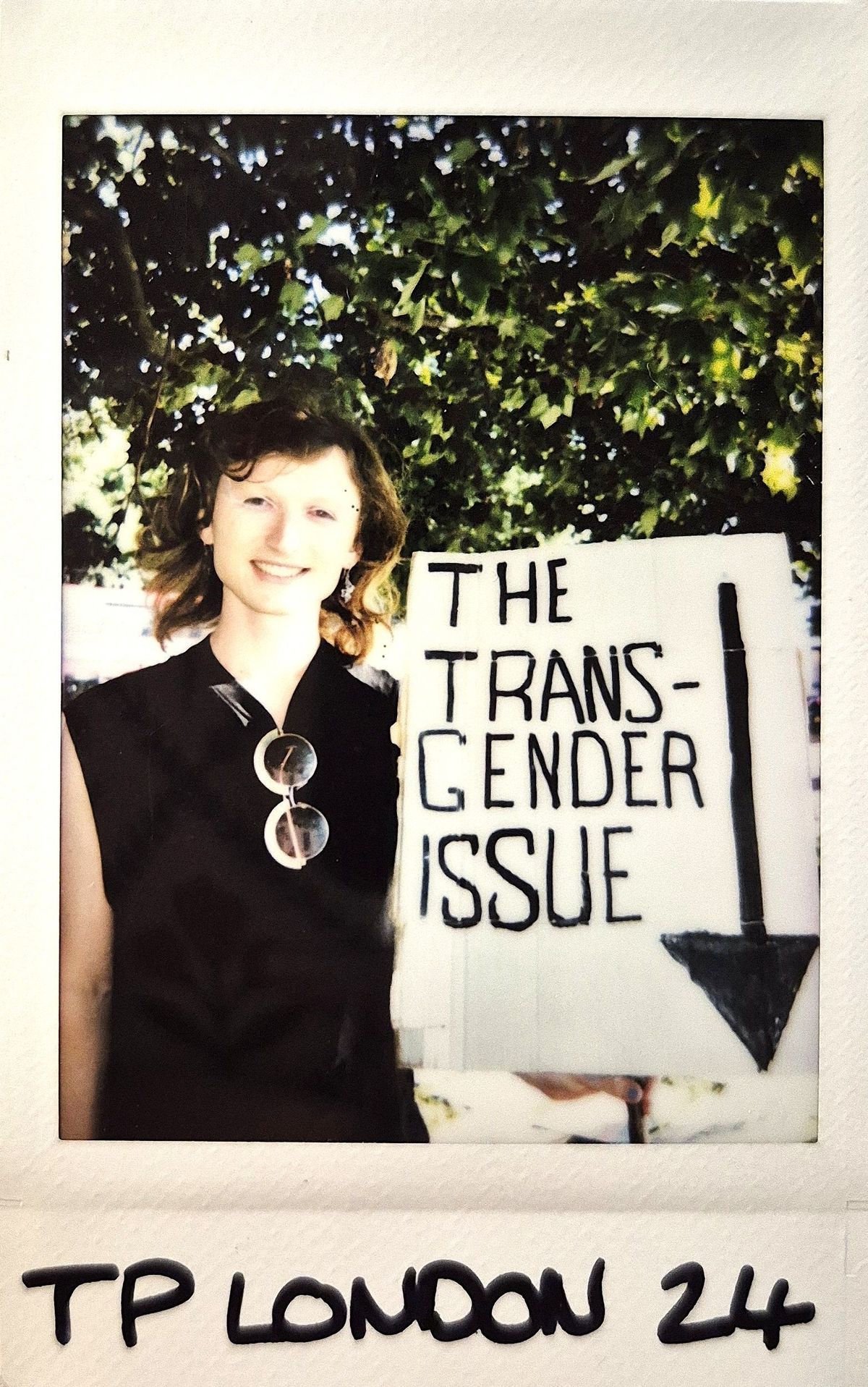 A smiling person stands beside a sign reading "The Transgender Issue" under a leafy tree canopy.