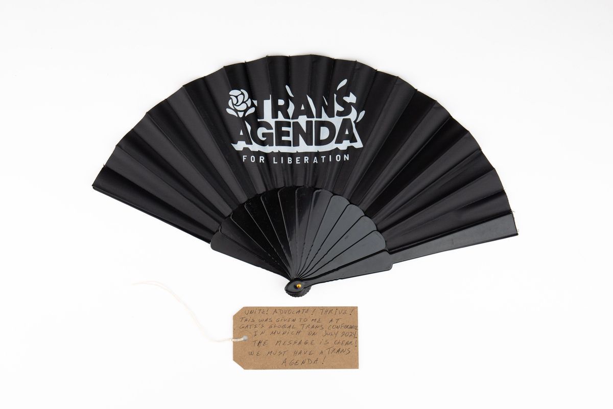 The image shows a black fan labeled "Trans Agenda for Liberation" with a handwritten tag.
