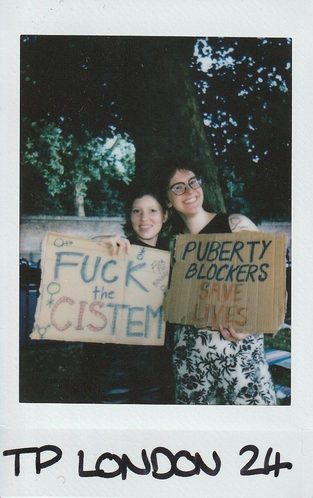 In the image, two individuals smilingly hold signs advocating for trans rights under a large tree outdoors.