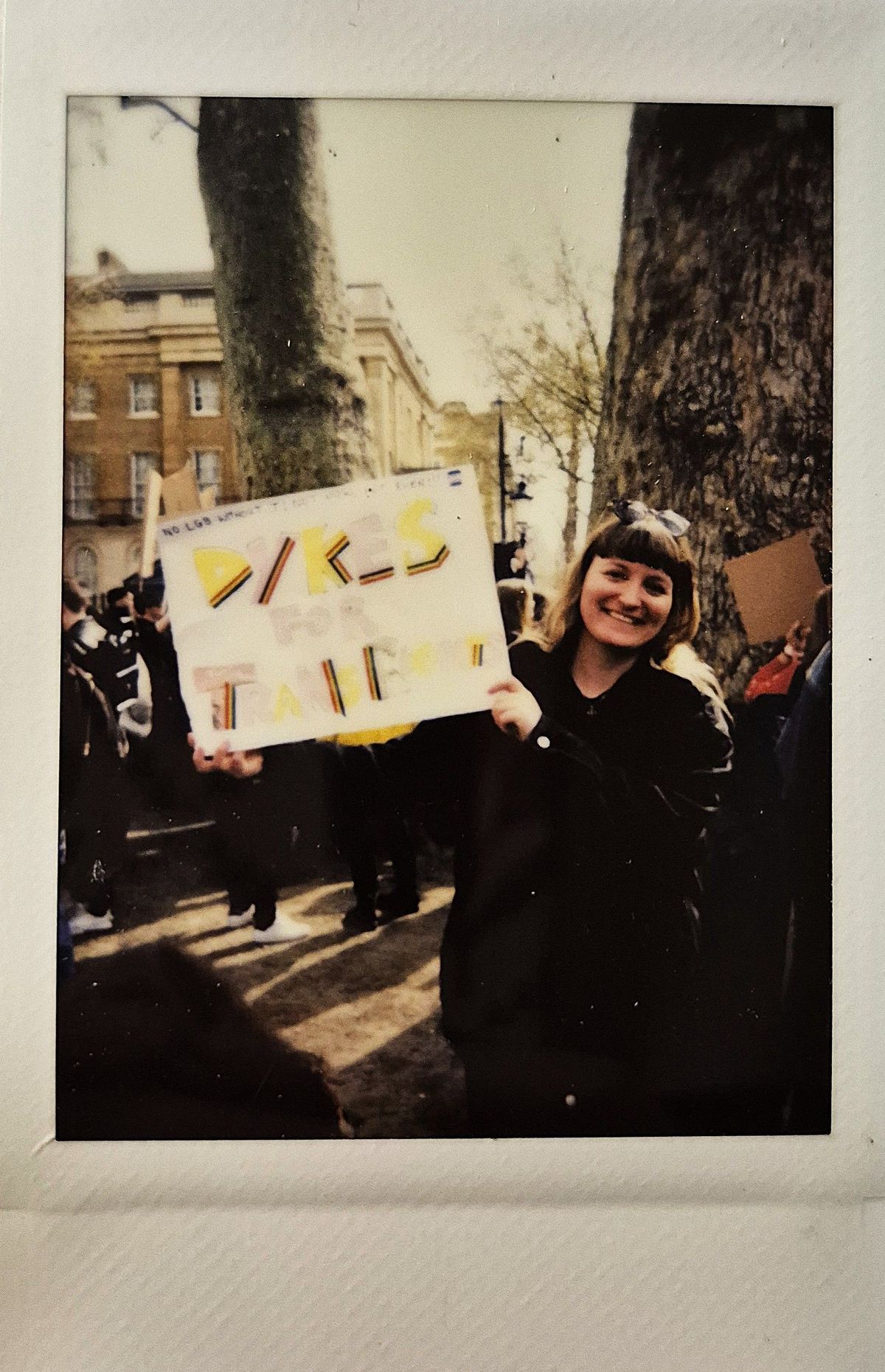 A person is joyfully holding a sign that says "Dykes for Trans" during an outdoor gathering.