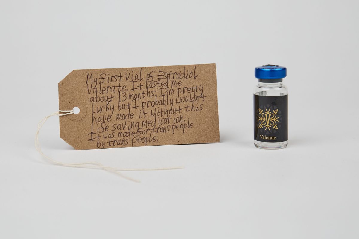 Brown luggage tag with vial of Estradiol Valerate, with a blue lid, on white background.