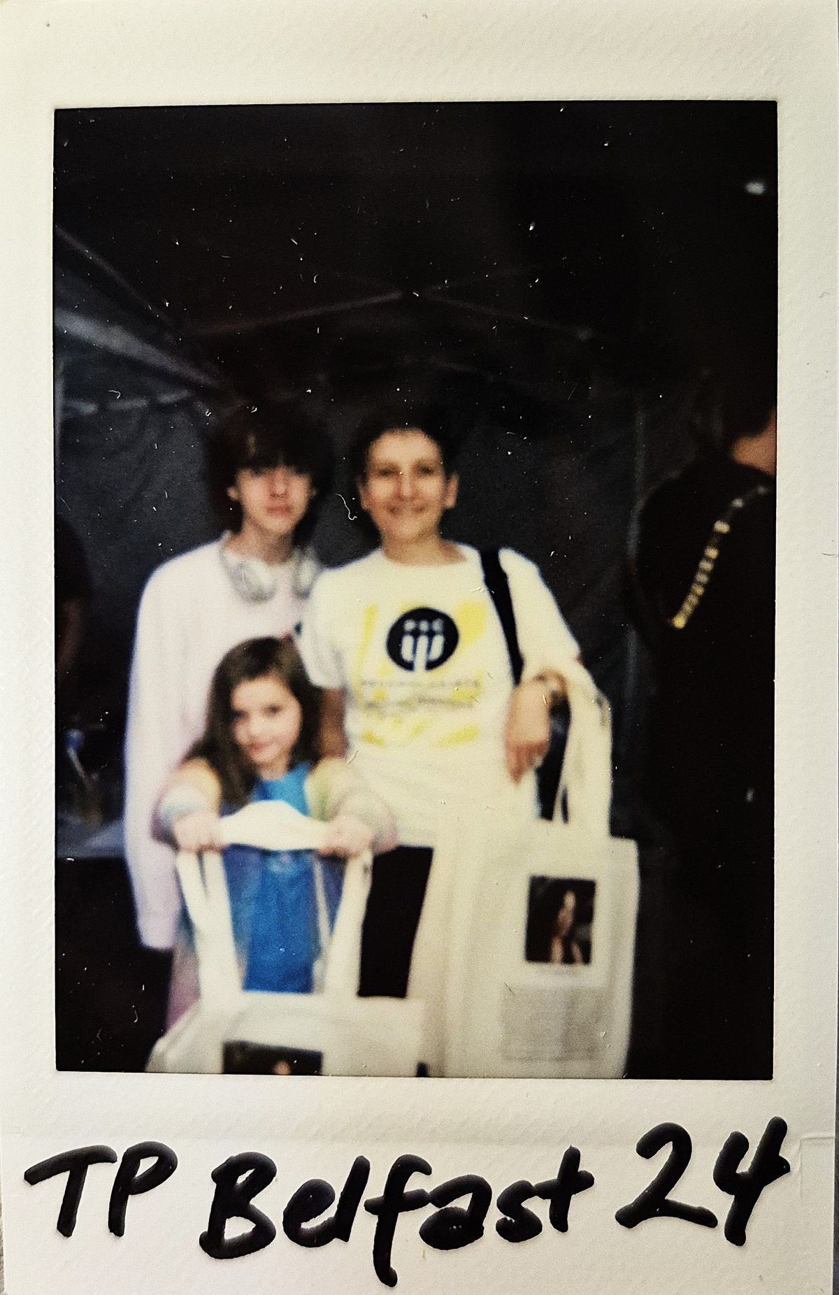 Three people are smiling for a Polaroid photo, labeled "TP Belfast 24".