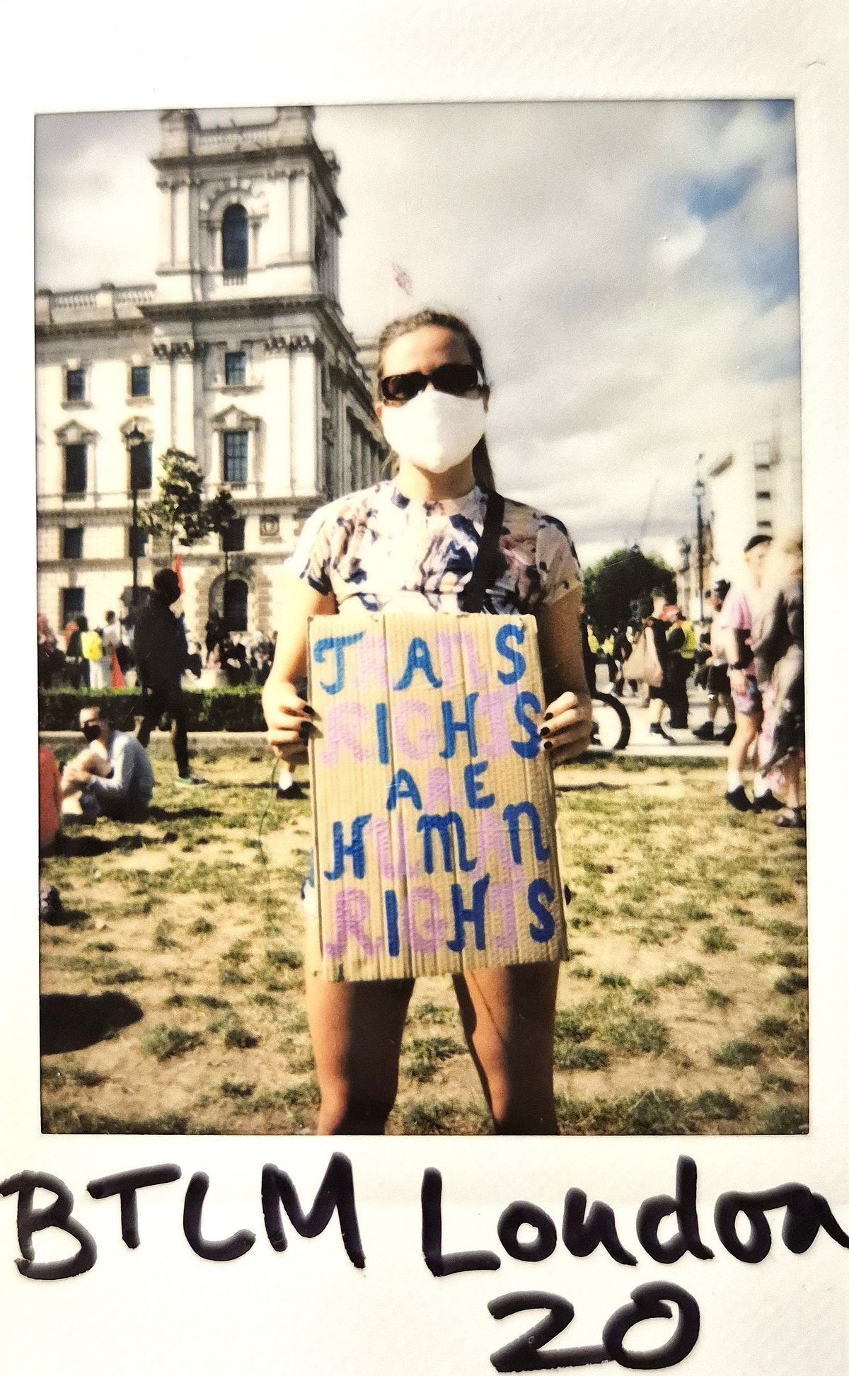 A person with sunglasses and mask holds sign for trans rights in a busy London square.