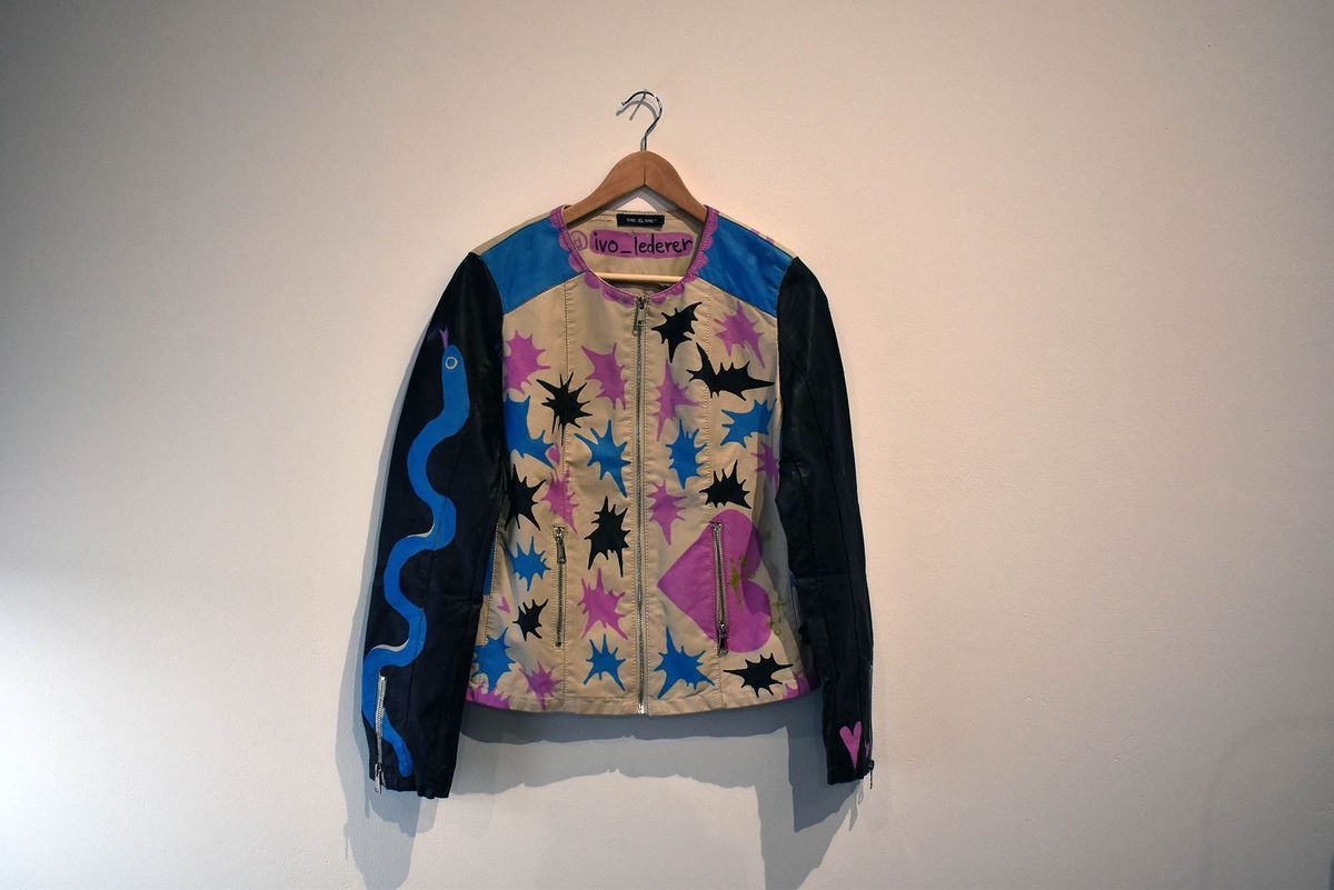 Front of leather jacket. The vest portion is white with purple sleeves. There are several paintings across the jacket - on the white front are splashes of colour. On the right arm is a blue painted snake, on the left arm a trans pride flag and pink hearts.