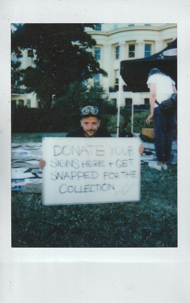 E-J Scott holds a placard which says "DONATE YOUR SIGNS HERE + GET SNAPPED FOR THE COLLECTION". Behind E-J is a grass field full of placard on the floor.
