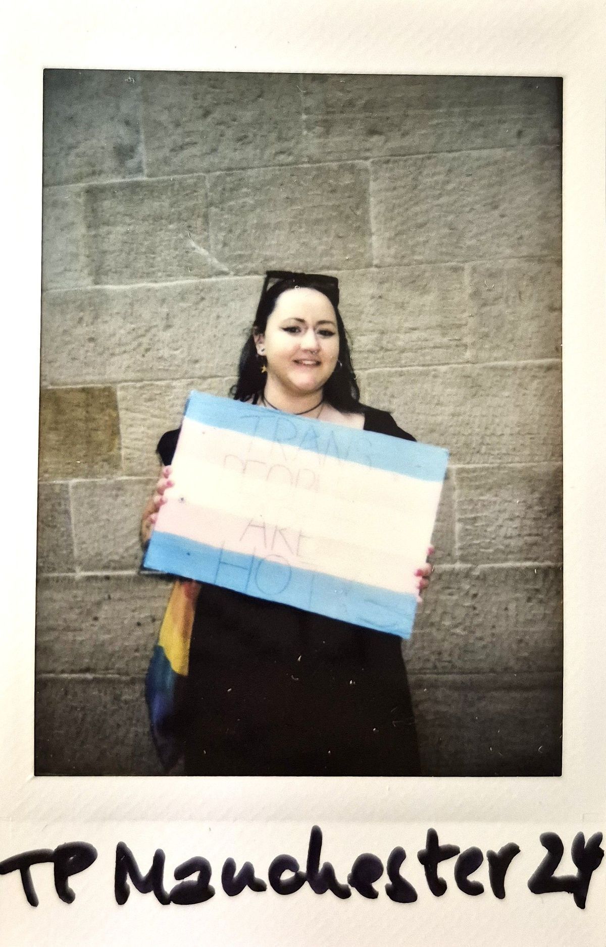 A person holds a sign with the transgender flag colors, standing against a stone wall.