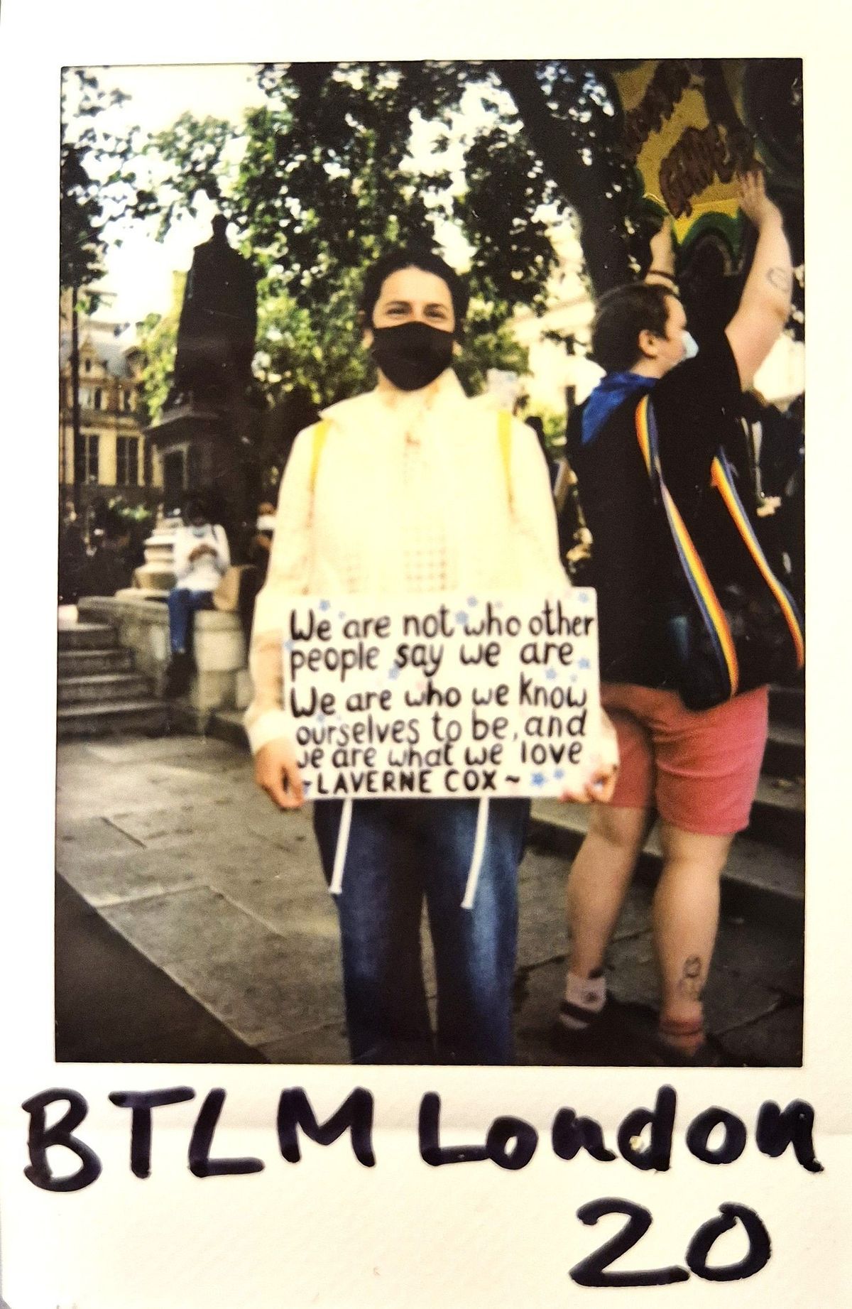 A person stands holding an inspirational sign by Laverne Cox, wearing a mask, in a lively