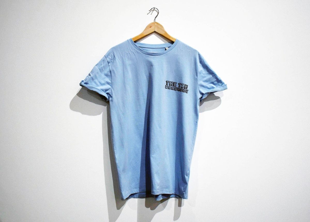 A light blue t-shirt with "THE TIN WARDROBE" printed on the breast on a wooden hanger.