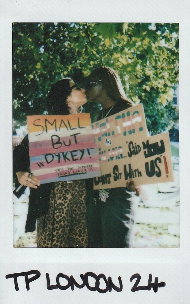 Two individuals are sharing a kiss while holding colorful protest signs under a tree.