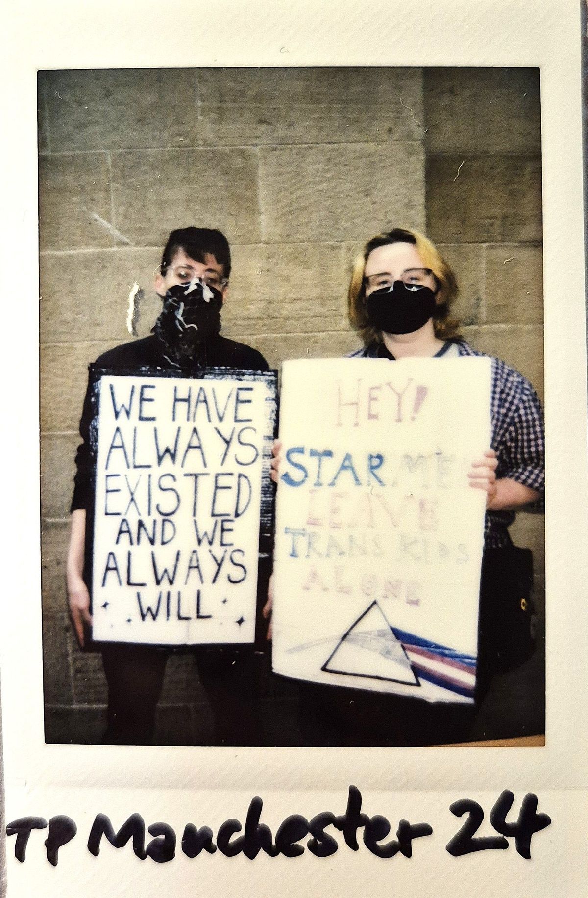 Two masked individuals hold protest signs advocating for trans rights, standing against a brick wall backdrop.