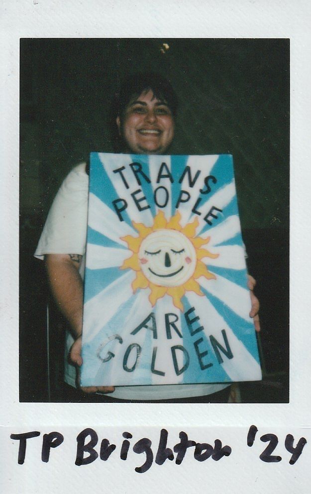 A person smiles, holding a colorful sign reading "Trans People Are Golden" with a sun illustration.