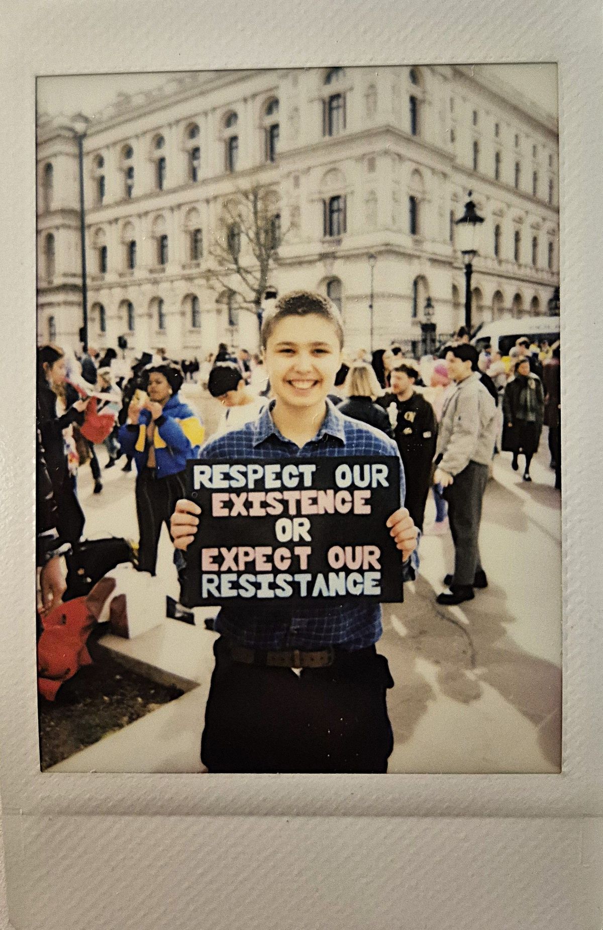 A person is holding a sign reading, "Respect our existence or expect our resistance," with a crowd behind them.