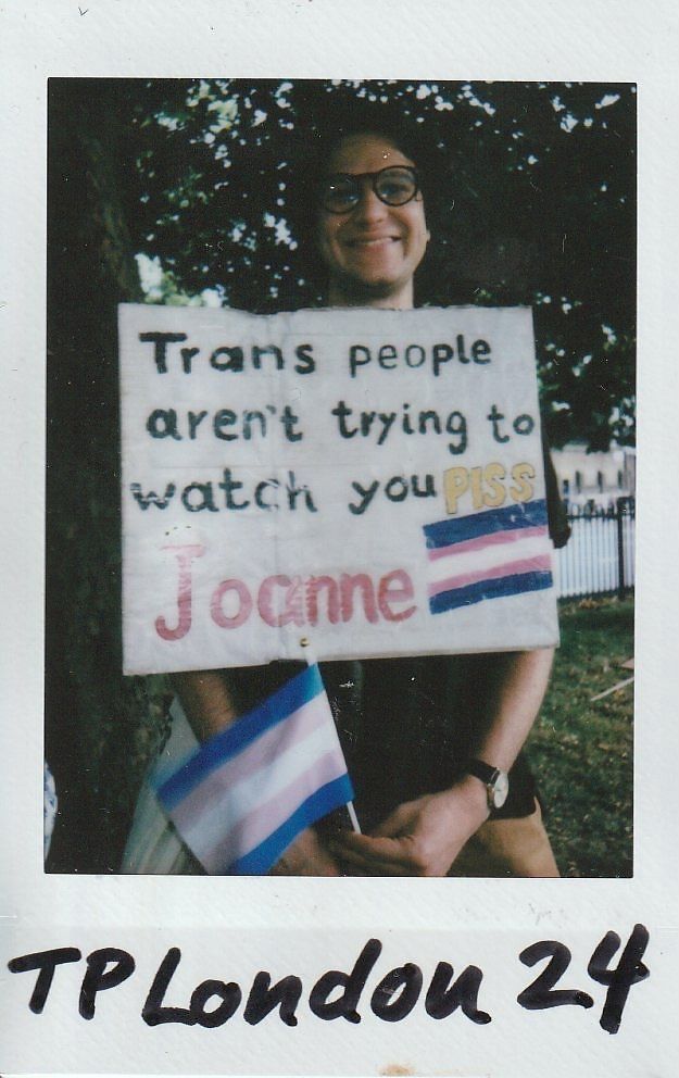 A person holds a sign which says "Trans People Aren't Trying To Watch You Piss Joanne" whilst they smile under a tree in a park setting.