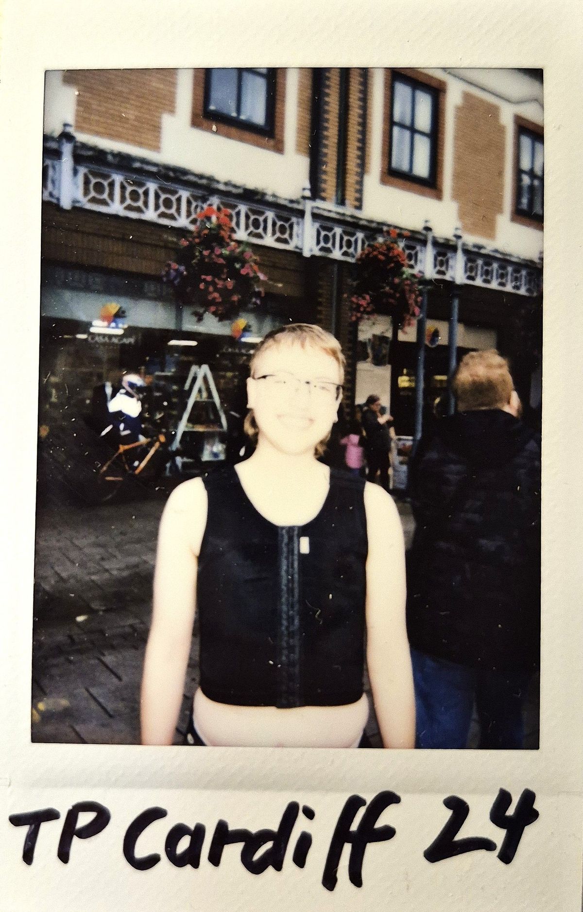 A person is smiling outdoors, wearing glasses and a black top, with buildings and people in the background.