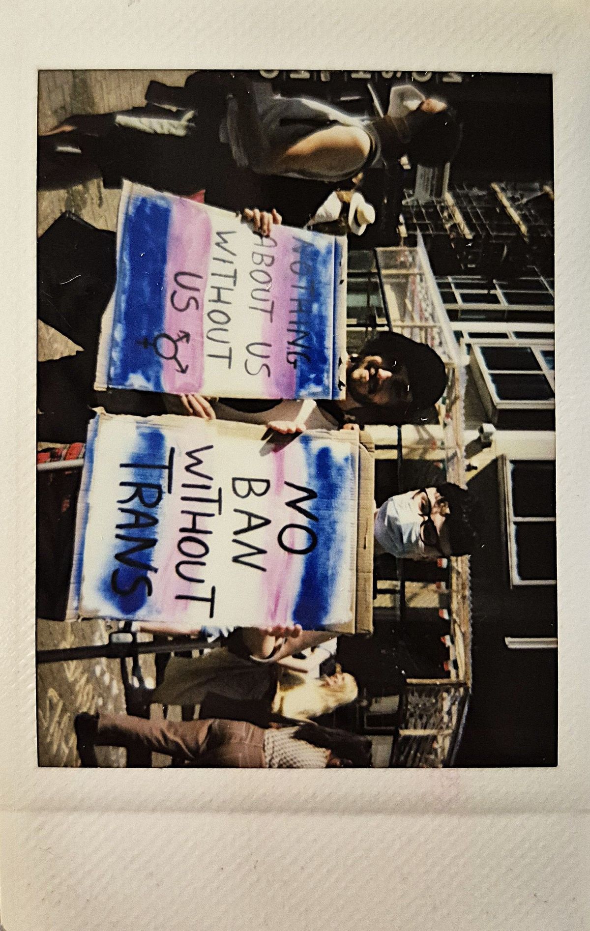 Two individuals hold signs which say "NOTHING ABOUT US WITHOUT US" and "NO BAN WITHOUT TRANS".