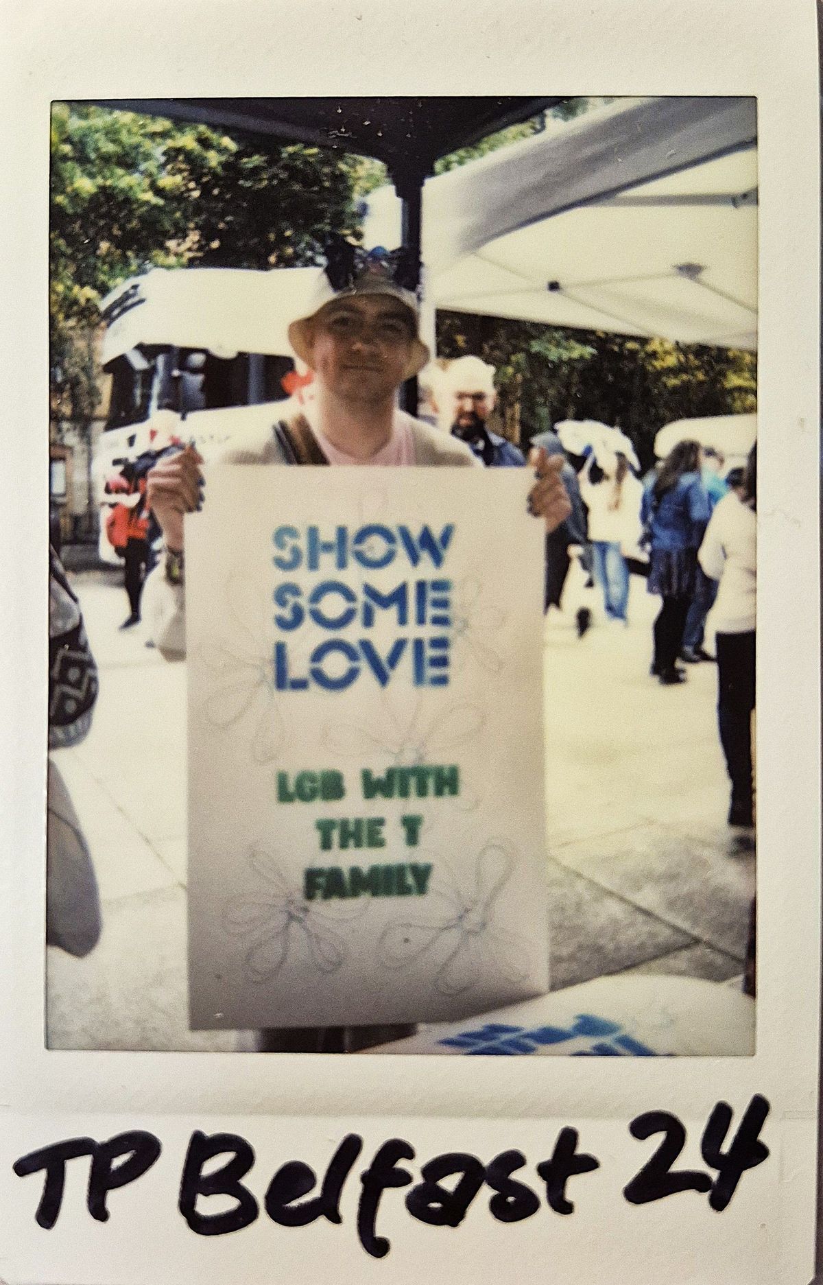 A person holds a sign saying “SHOW SOME LOVE LGB WITH THE T FAMILY” at an outdoor event.