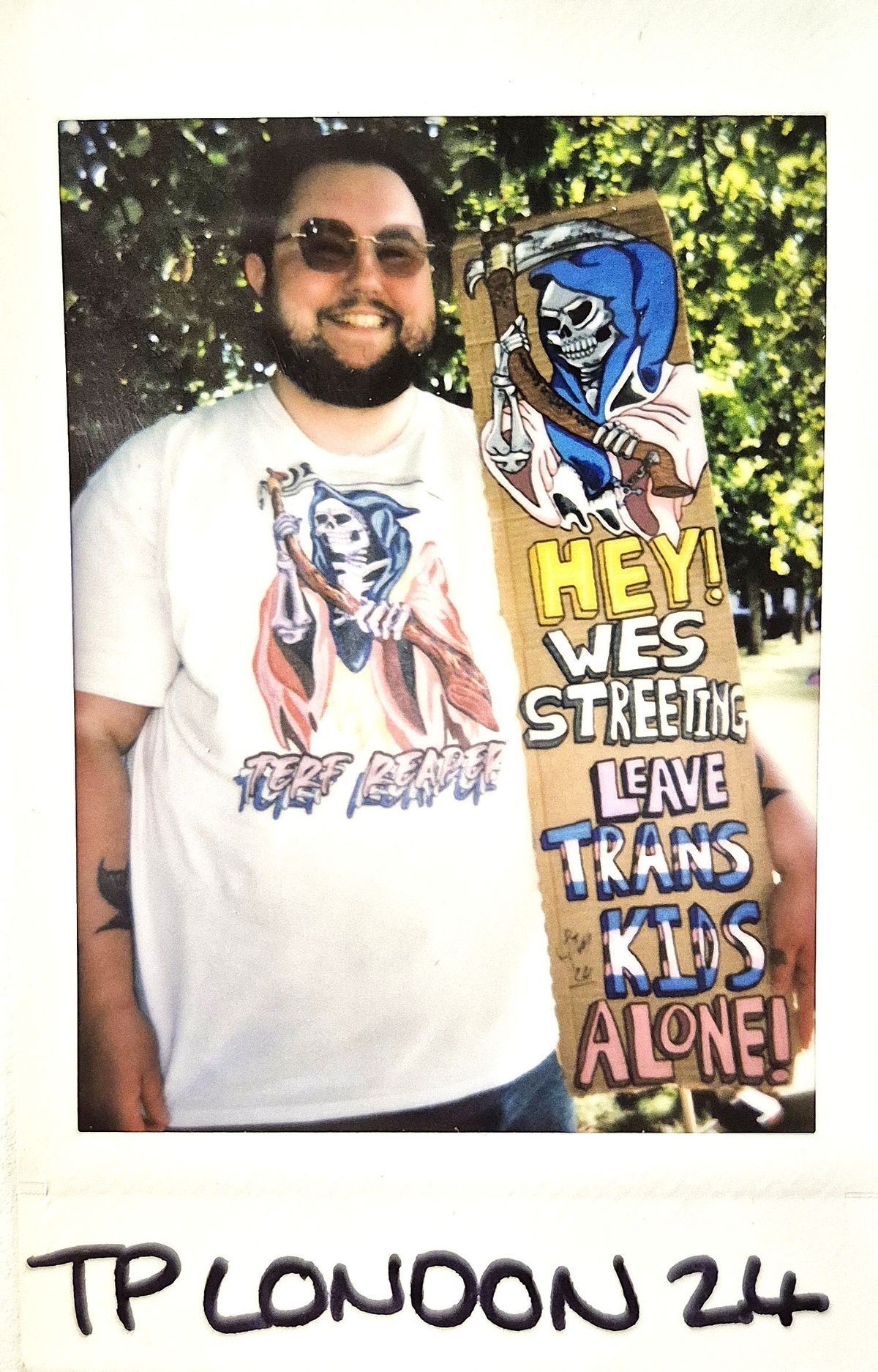 A person holds a colorful sign advocating for trans kids, wearing matching themed sunglasses and shirt.