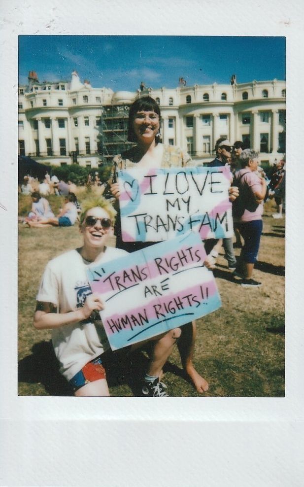 Two individuals in the sun smiling and holding two placards, one says "I Love My Trans Fam" and the other says "Trans Rights Are Human Rights".