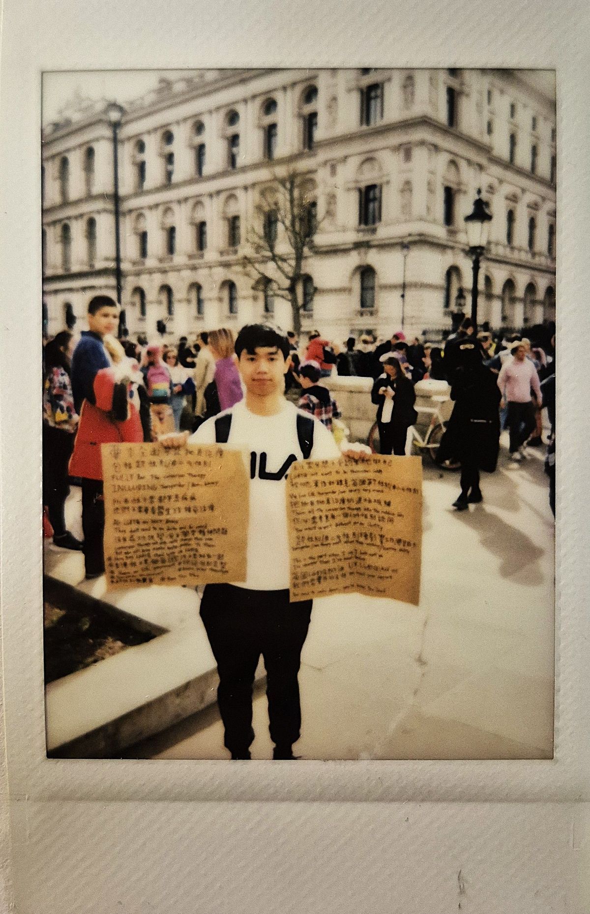A person holds two written signs in a crowded public square, with a historic building behind them.