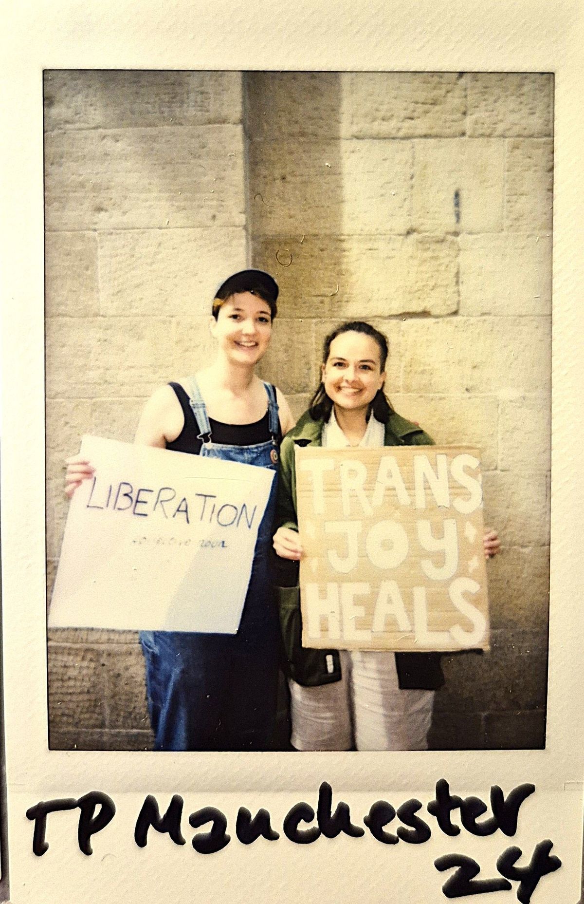 Two people are holding signs saying “LIBERATION” and “TRANS JOY HEALS”, smiling against a wall.