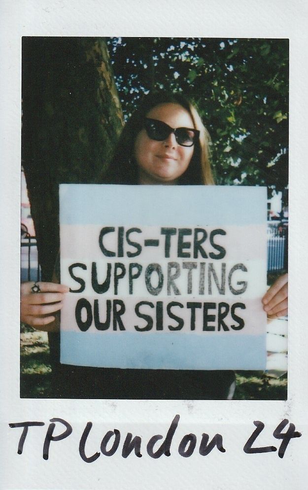 A person wearing sunglasses holds a sign that reads "Cis-ters Supporting Our Sisters," standing outdoors