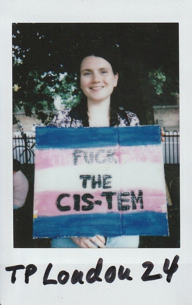 A person holds a colorful protest sign with a message challenging societal norms in a park setting.