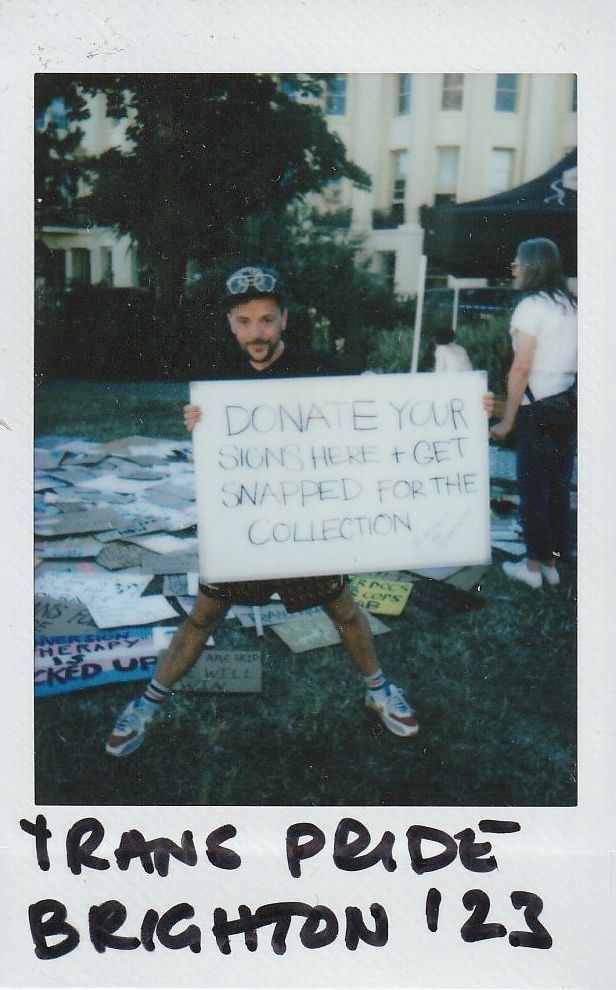 E-J Scott holds a placard which says "DONATE YOUR SIGNS HERE + GET SNAPPED FOR THE COLLECTION". Behind E-J is a grass field full of placard on the floor.