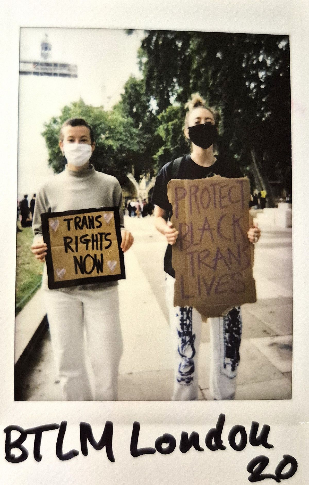 Two individuals, wearing masks and holding protest signs, stand in support of trans rights and equality.