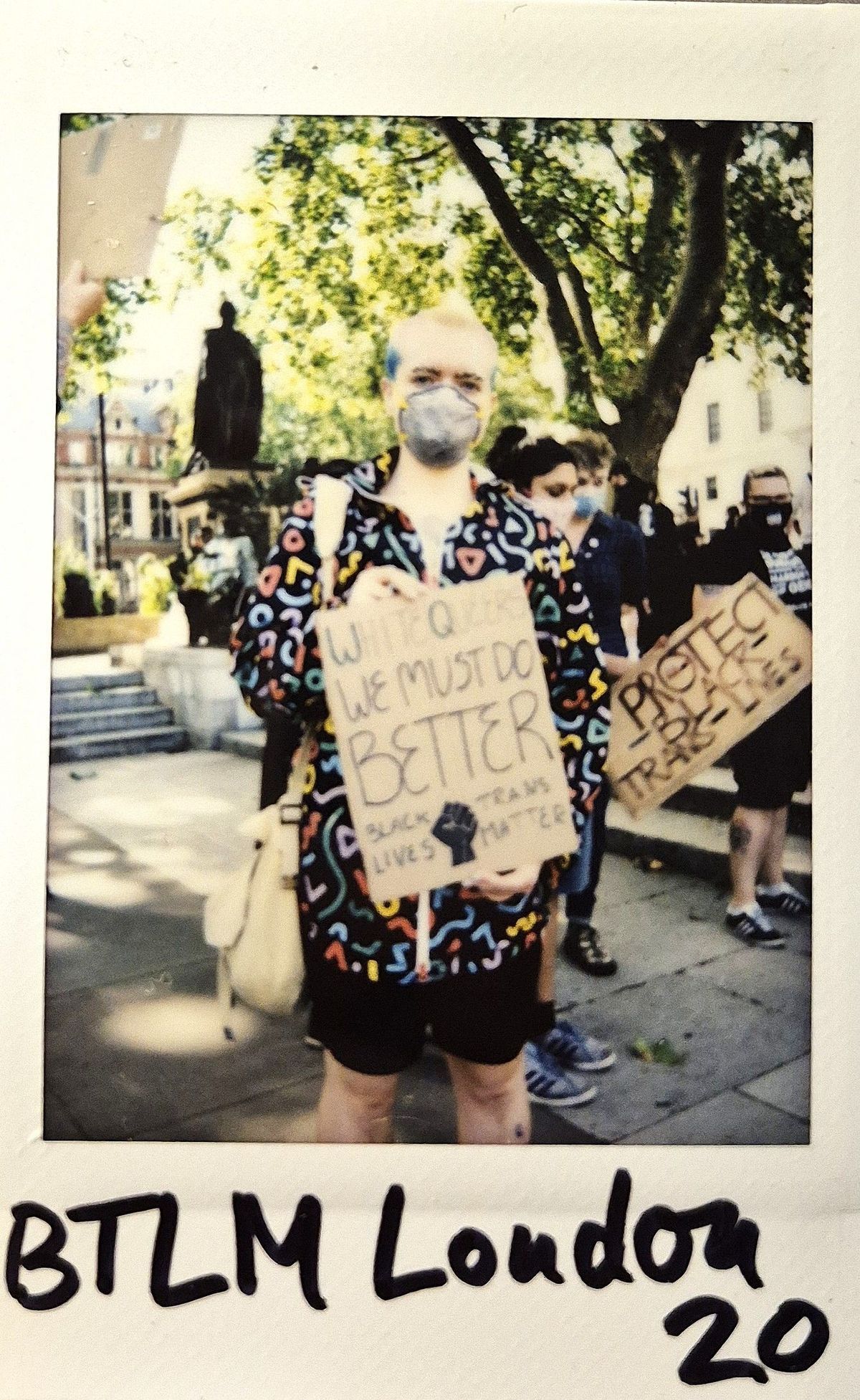 A person stands in a colorful hoodie, holding a protest sign amidst a group of people in a park