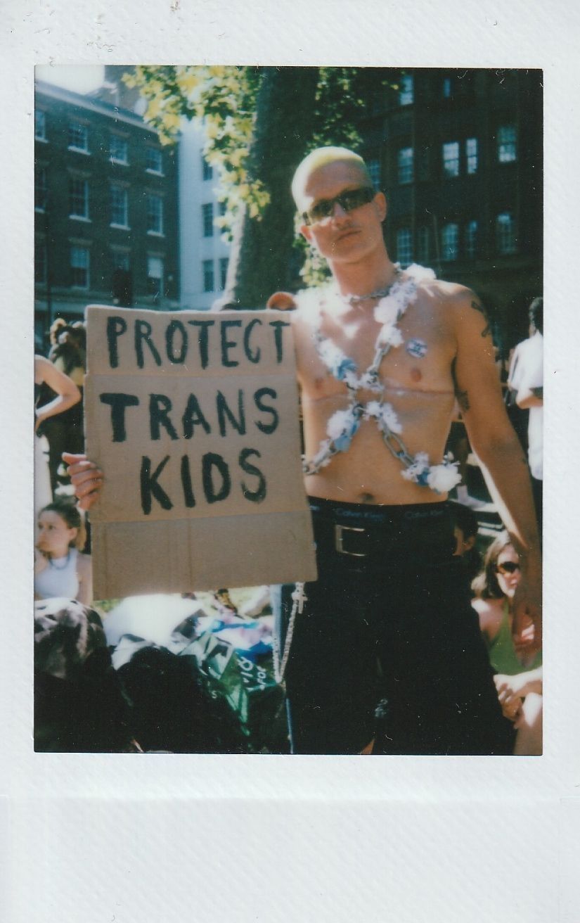 A person stands outdoors holding a cardboard sign that reads "Protect Trans Kids" during a sunny day.