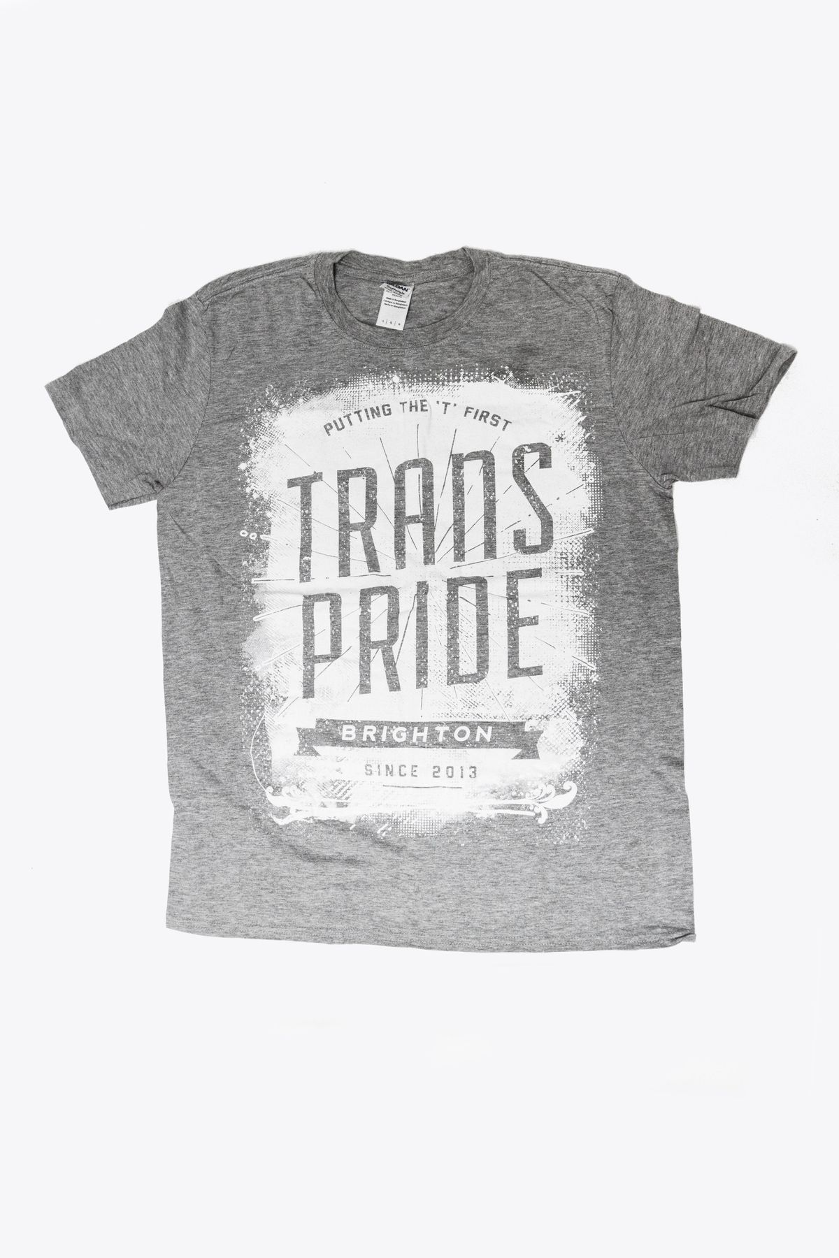 A grey T-shirt with the text "Putting the 'T' First" at the top, "Trans Pride" in large letters in the middle "Brighton" on a banner underneath, and "Since 2013" at the bottom. A label with the brand "Gildan" can be seen on the neck.