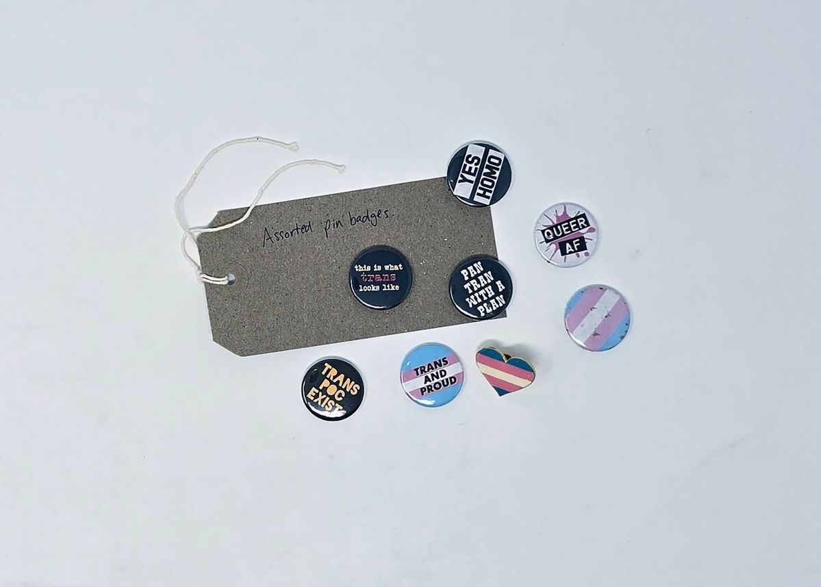 Assorted pin badges with various pride slogans and designs are displayed on a white surface.
