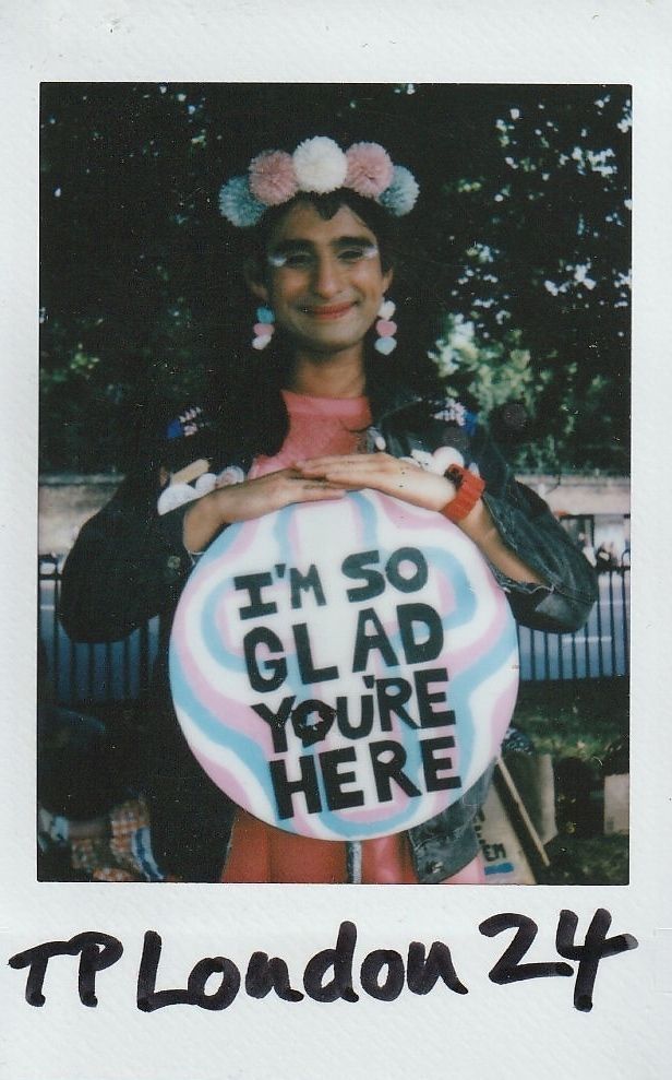 A person with colorful accessories holds a sign reading, "I'M SO GLAD YOU'RE HERE".