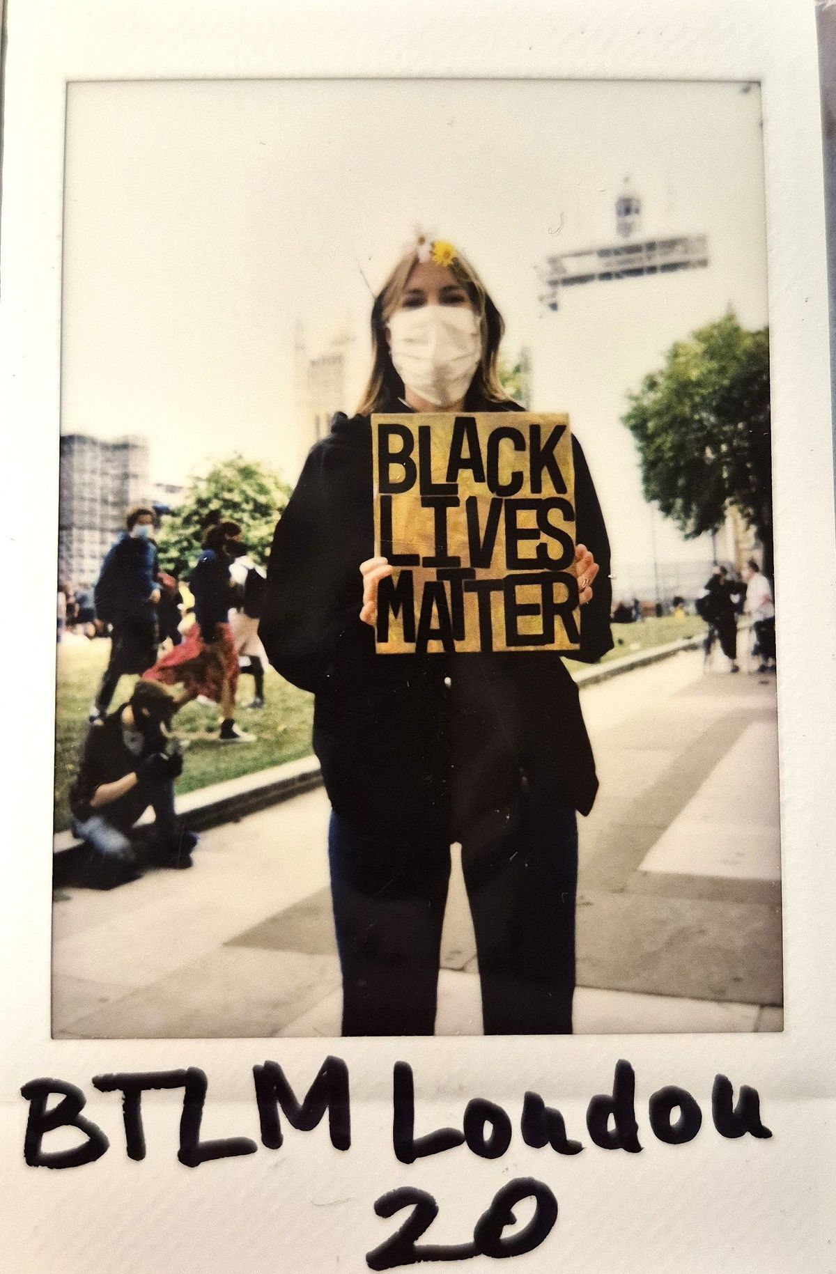 A masked individual holds a "Black Lives Matter" sign during a public demonstration outdoors with others nearby.