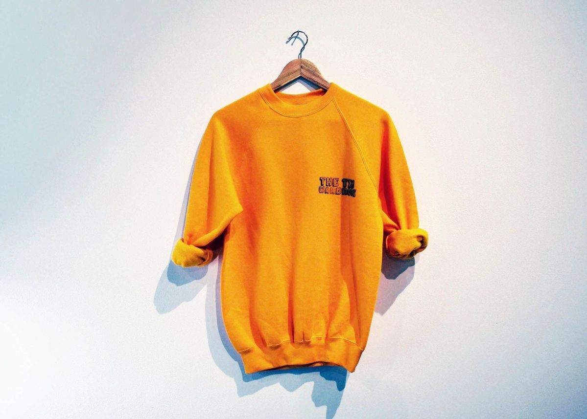 A yellow sweatshirt with rolled up sleeves and "THE TIN WARDROBE" printed on the breast, on a wooden hanger.