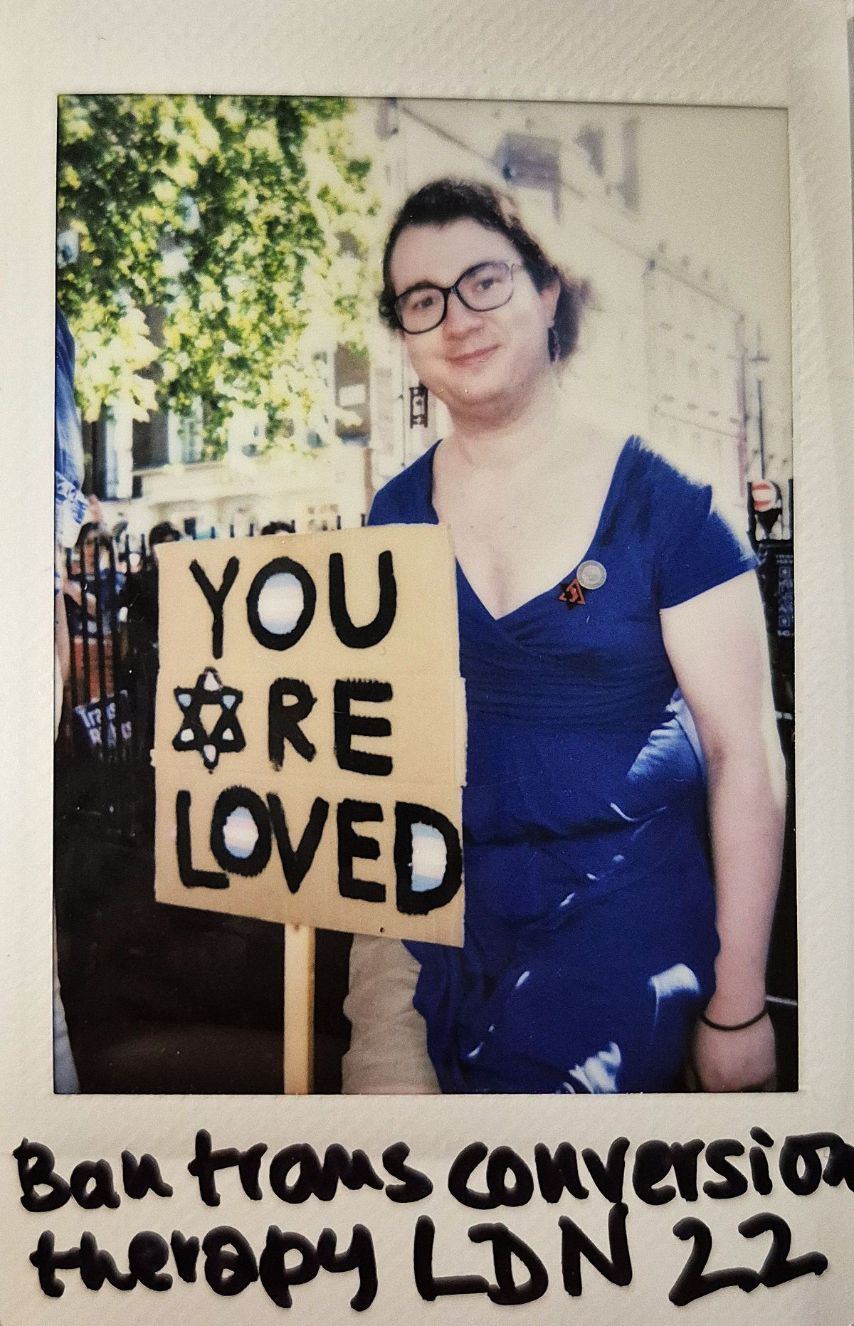 A person in a blue dress holds a sign reading "YOU ARE LOVED" at a protest.