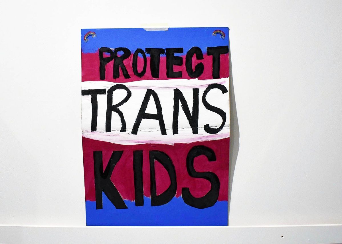 The image shows a colorful sign stating "Protect Trans Kids" in bold letters.