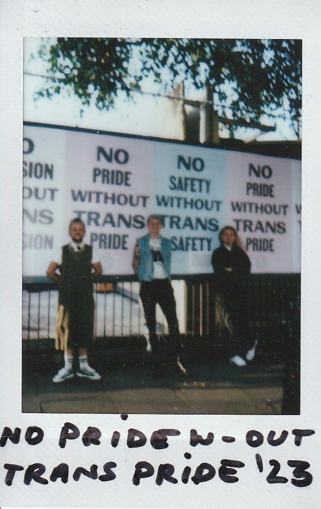 Three people stand in front of signs which say "No Pride Without Trans Pride".